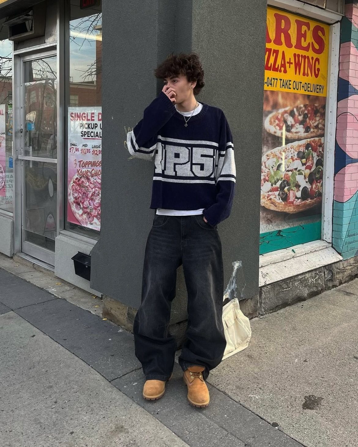 A young man posing in a navy sweater and wide-leg black trousers on a city street.
