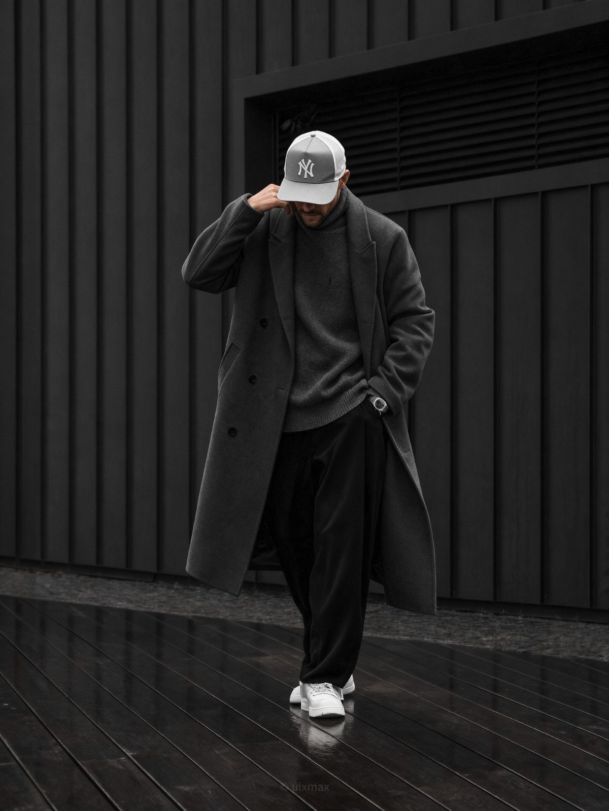 Man in a long grey coat, grey turtleneck, and black trousers standing against a dark wall.