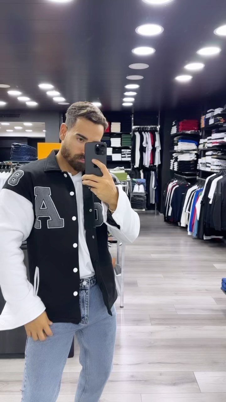 Man in a black and white varsity jacket and light blue jeans taking a mirror selfie.