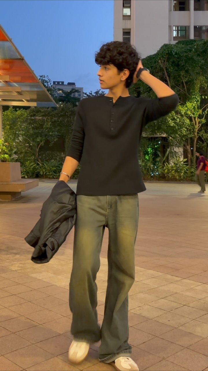 A young man posing in a black henley shirt and olive green wide-leg trousers outdoors.