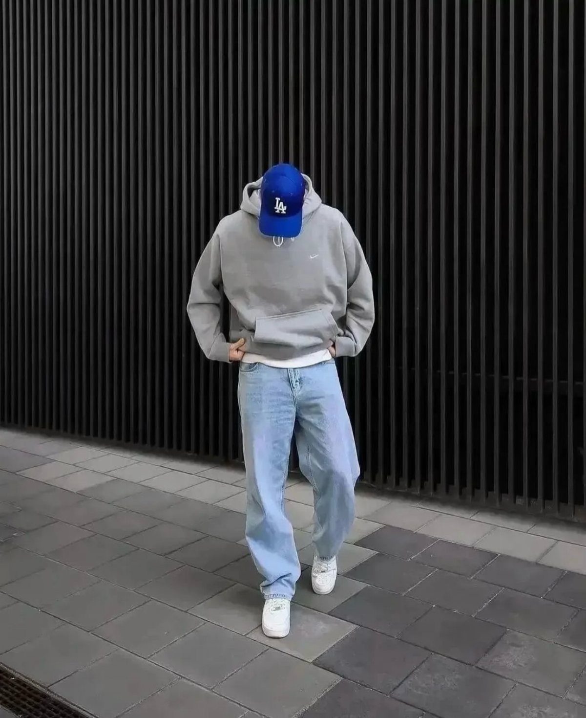 Person in a grey hoodie, baggy light blue jeans, and a blue cap standing against a black wall.