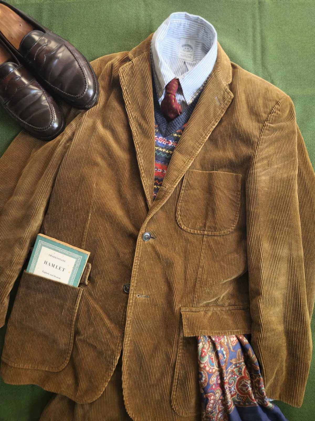 Brown corduroy blazer, striped shirt, red tie, and leather loafers arranged on a green background.