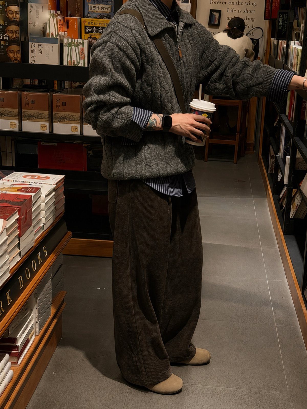 Person wearing a grey cable knit sweater and brown corduroy trousers in a bookstore.