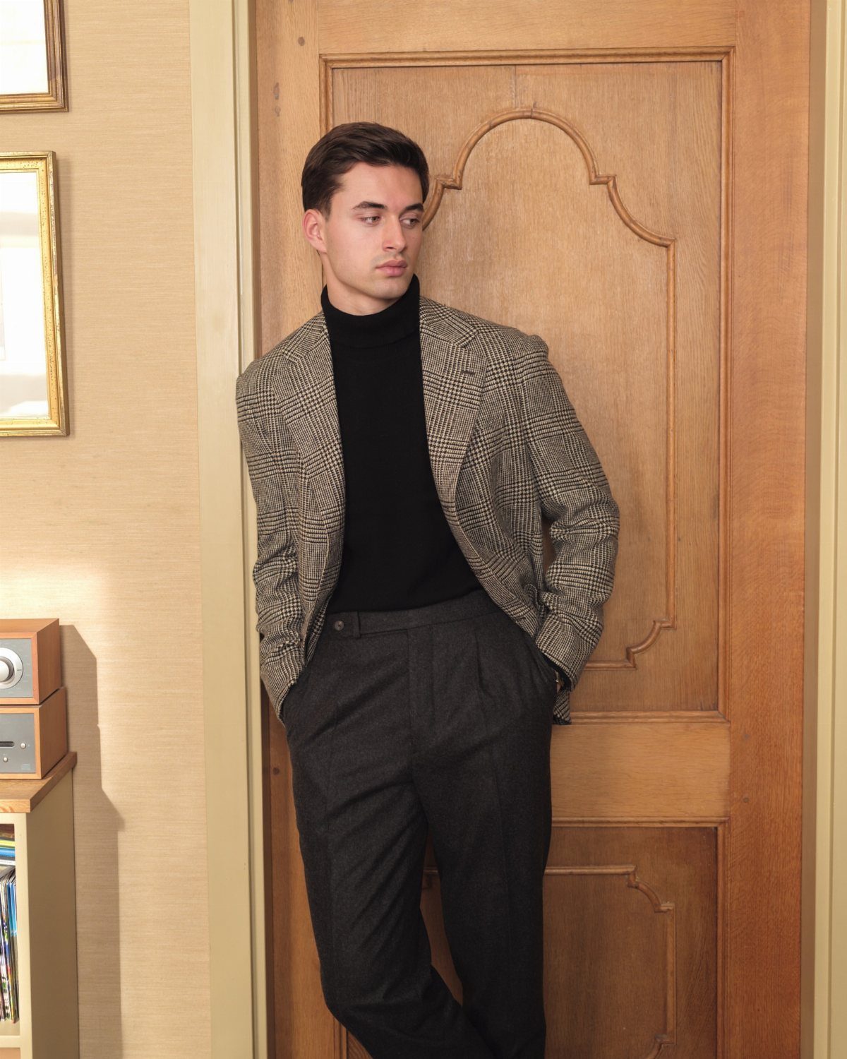 Man wearing a glen plaid oversized blazer, black turtleneck, and dark trousers leaning against a door.