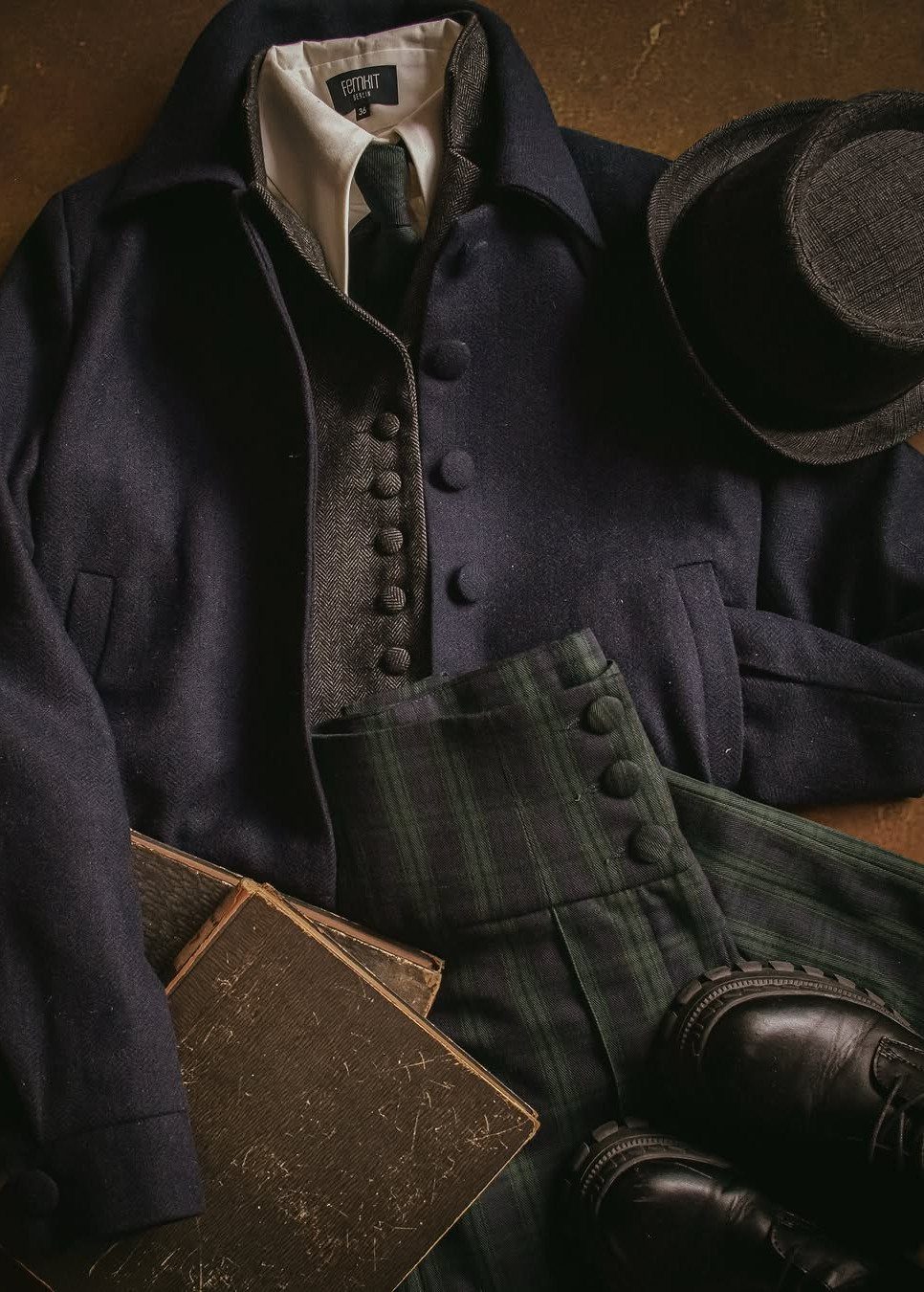 Dark academia clothing items including a navy coat, plaid trousers, waistcoat, and old books laid out.