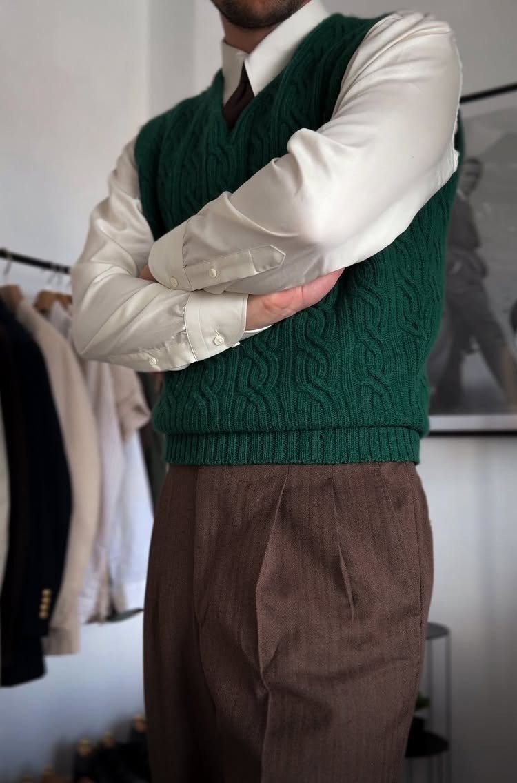 Man wearing a white shirt, green cable knit sweater vest, and brown pleated trousers.