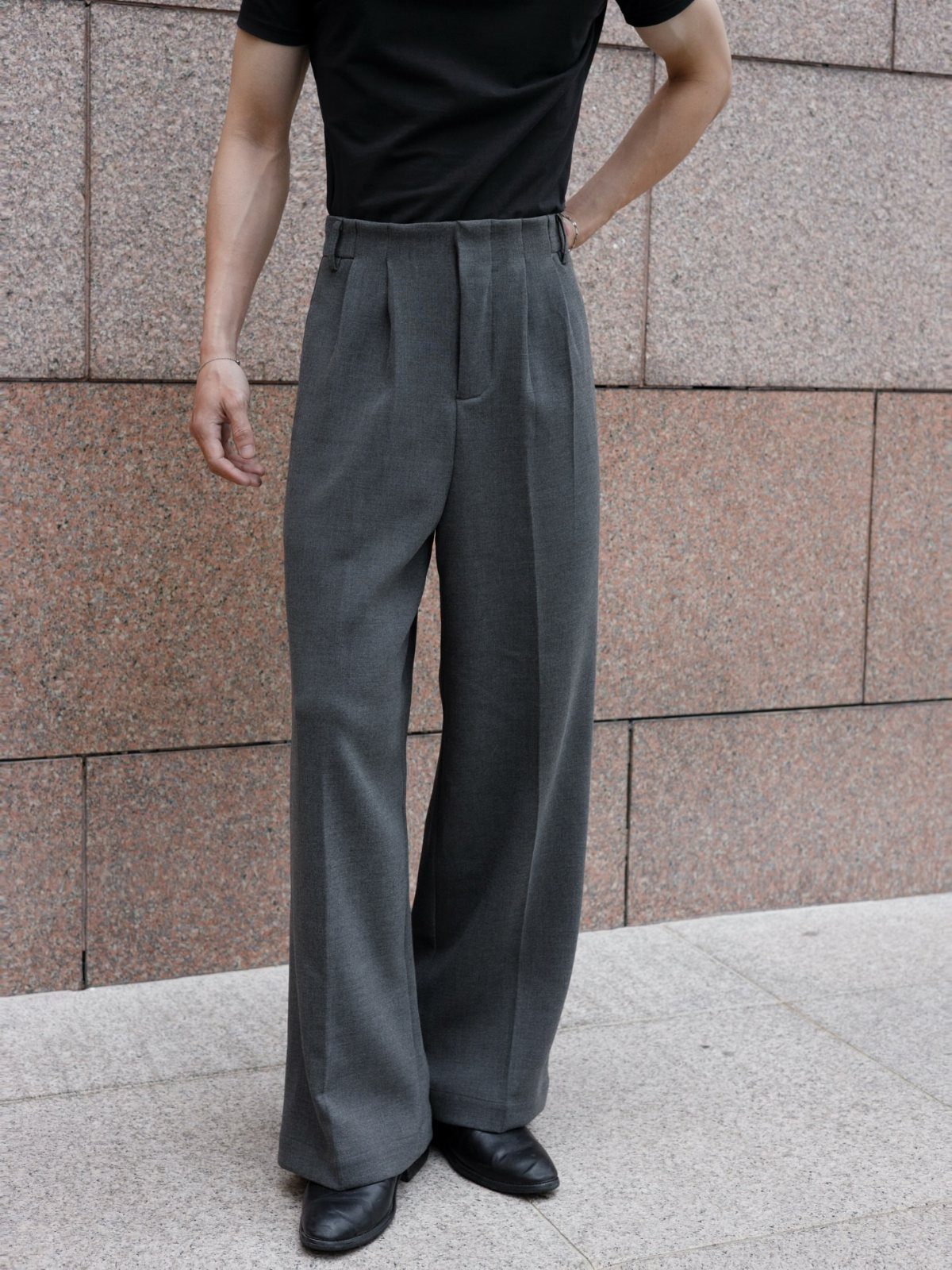 Person wearing wide-leg grey pleated tailored trousers and a black shirt against a stone wall.