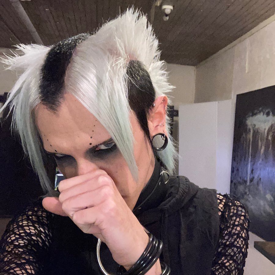 Person with spiky white and black hair, heavy dark eye makeup, and facial piercings.