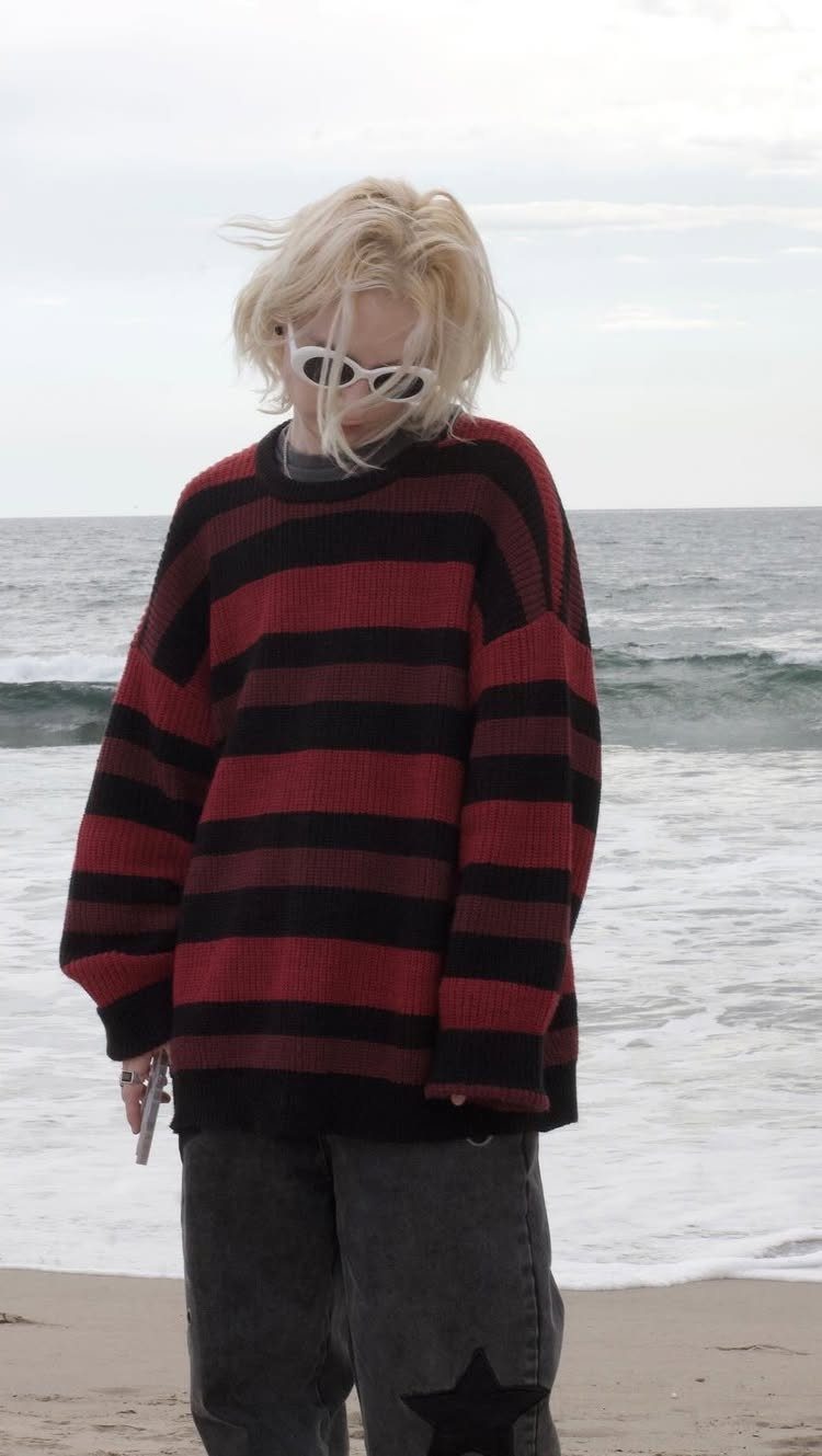 A person in a red and black striped oversized sweater standing on a sandy beach.