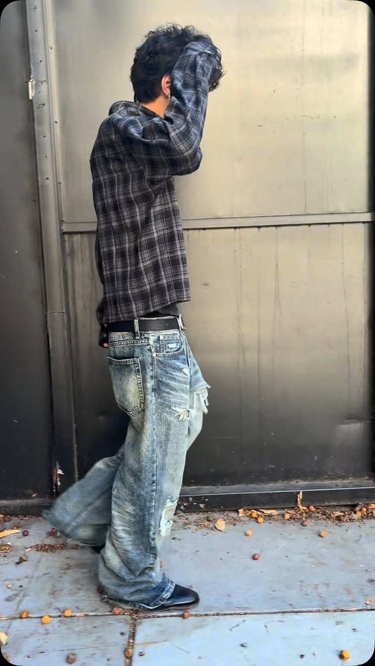 Person in a dark plaid flannel shirt and baggy ripped blue jeans standing against a black wall.