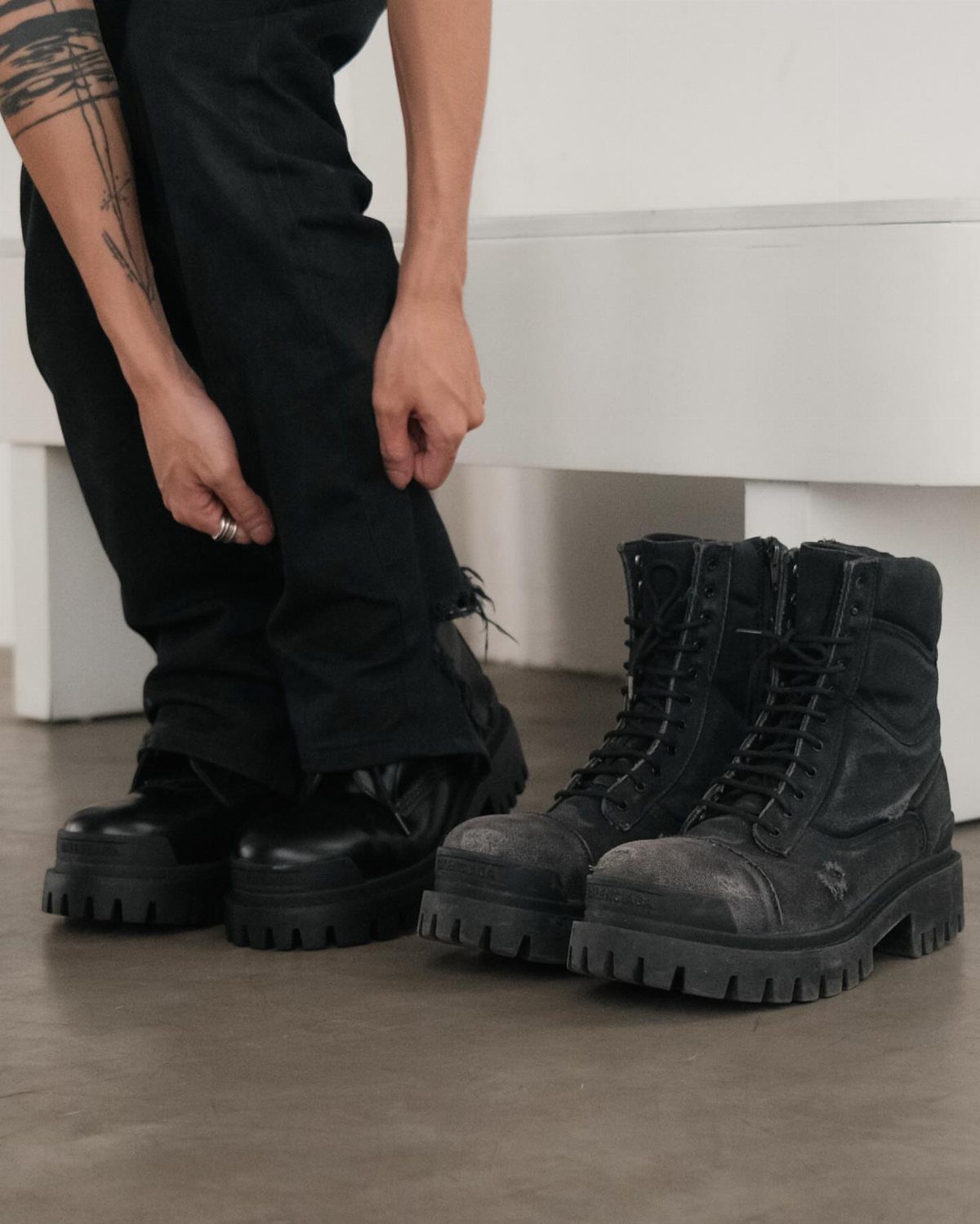 Black lace-up combat boots with thick platform soles sitting on a grey floor next to a person.