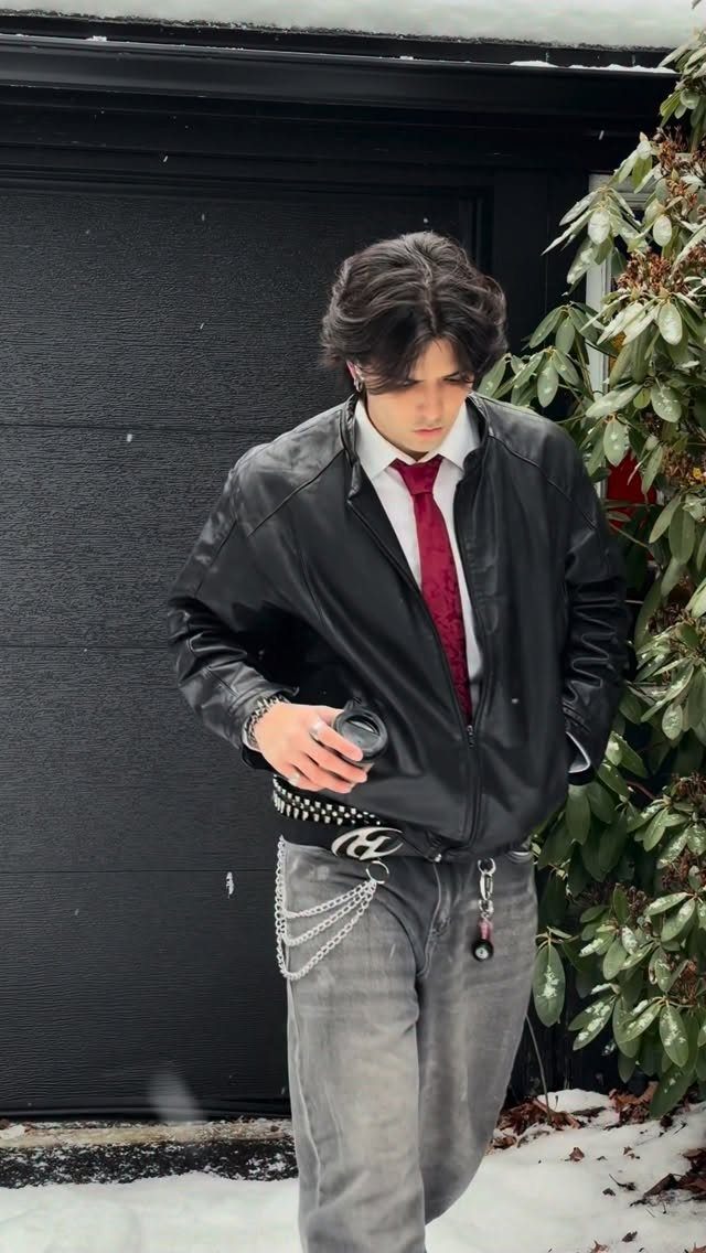 A person wearing a black leather jacket, red tie, and grey jeans with chains standing in snow.