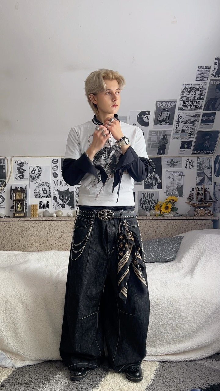 A person posing in a white and black layered outfit with wide-leg dark denim pants and silver chains.