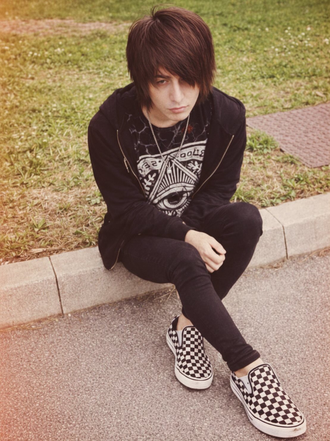 Young person in emo clothing sitting on a curb wearing a black hoodie and checkered shoes.