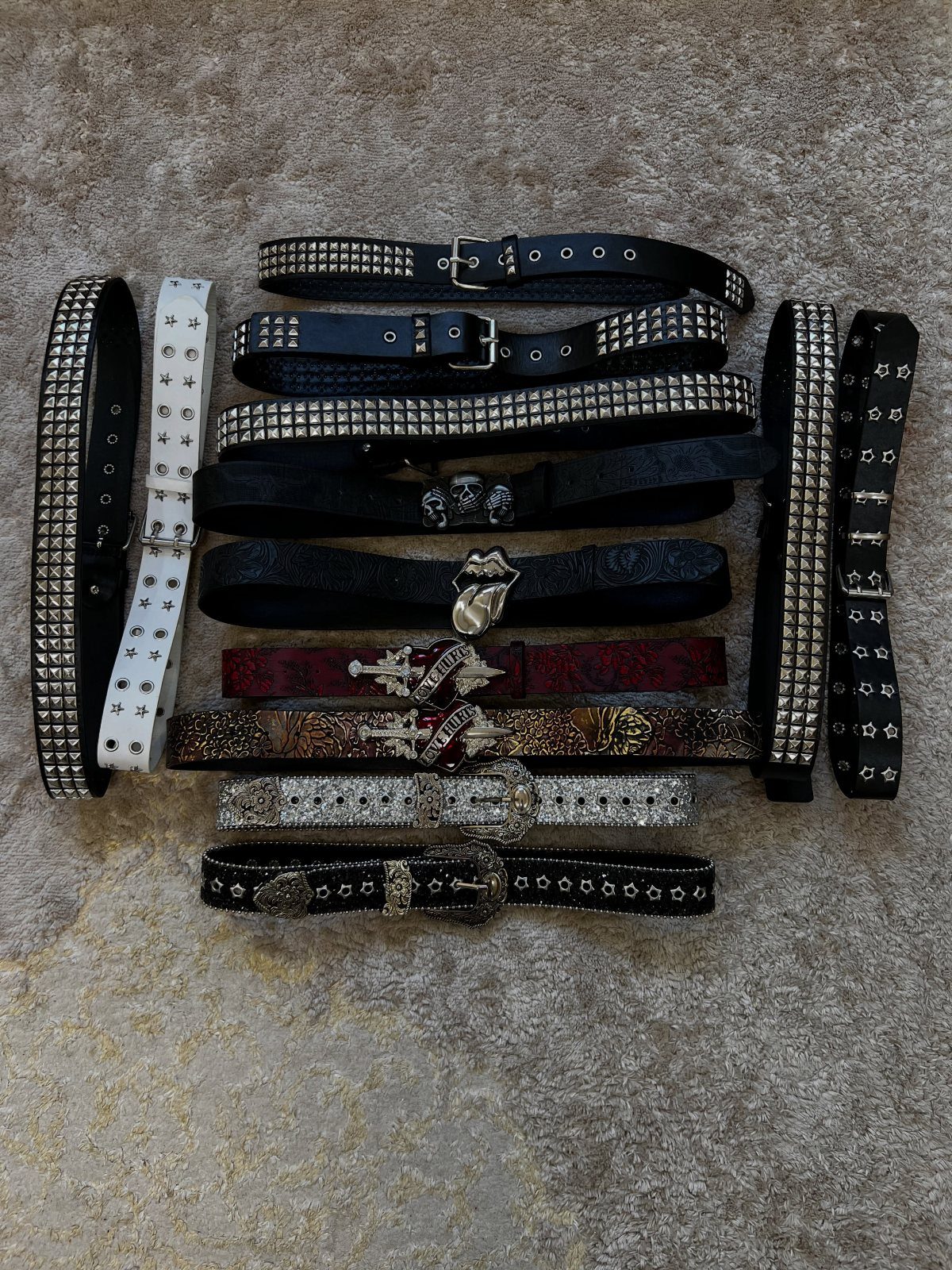 Multiple black and white studded leather belts with silver metal hardware laid on a beige carpet.