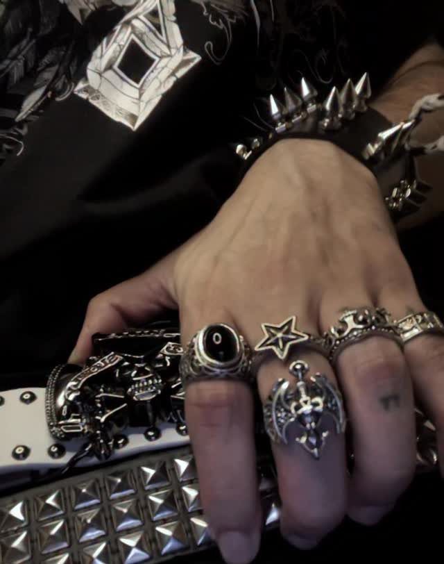 A hand wearing various silver gothic rings and a studded leather wristband holding a belt.
