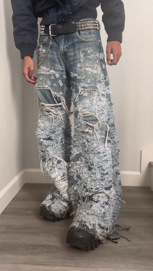 Person wearing heavily distressed, wide-leg light blue denim jeans with extreme fraying and large rips.