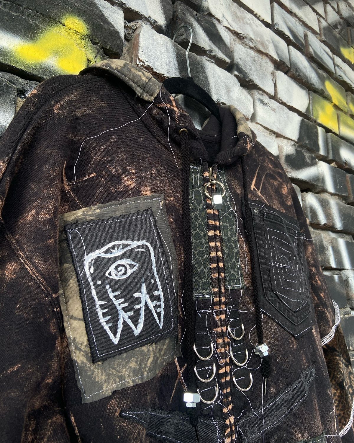 Distressed camouflage hoodie with patches and metal rings hanging against a graffiti brick wall.