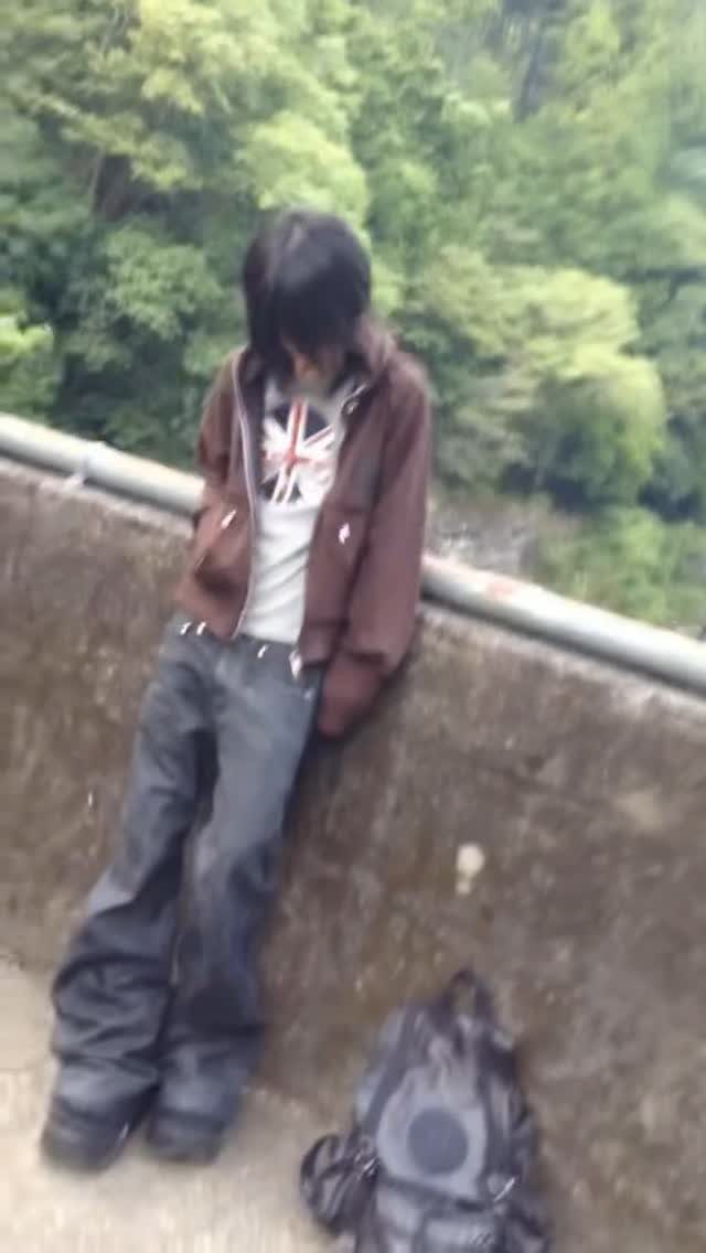 A person with dark hair wearing a brown hoodie and baggy jeans leans against a wall.