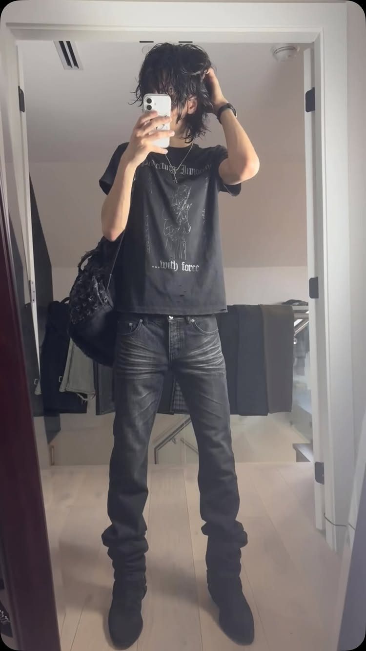 A person posing in a mirror wearing a black graphic t-shirt and dark skinny jeans.