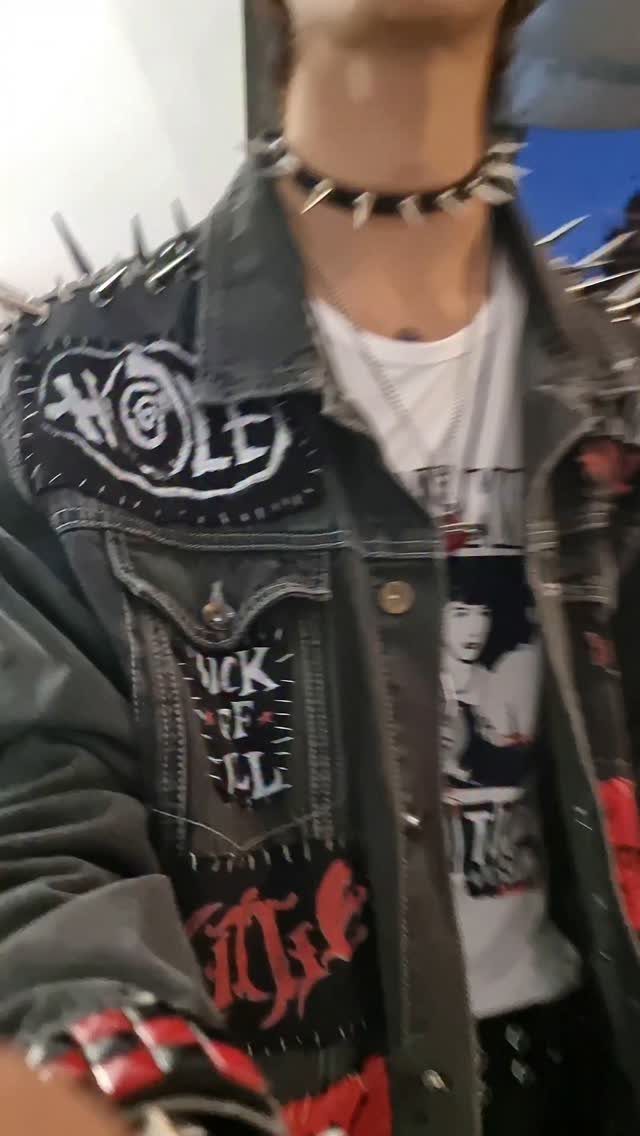 A person wearing a spiked choker and a patched, studded denim jacket over a graphic t-shirt.