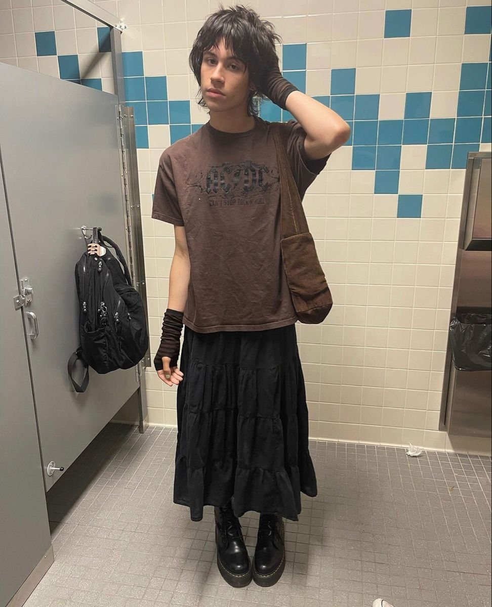 A person wearing a brown graphic t-shirt and a black tiered maxi skirt in a restroom.