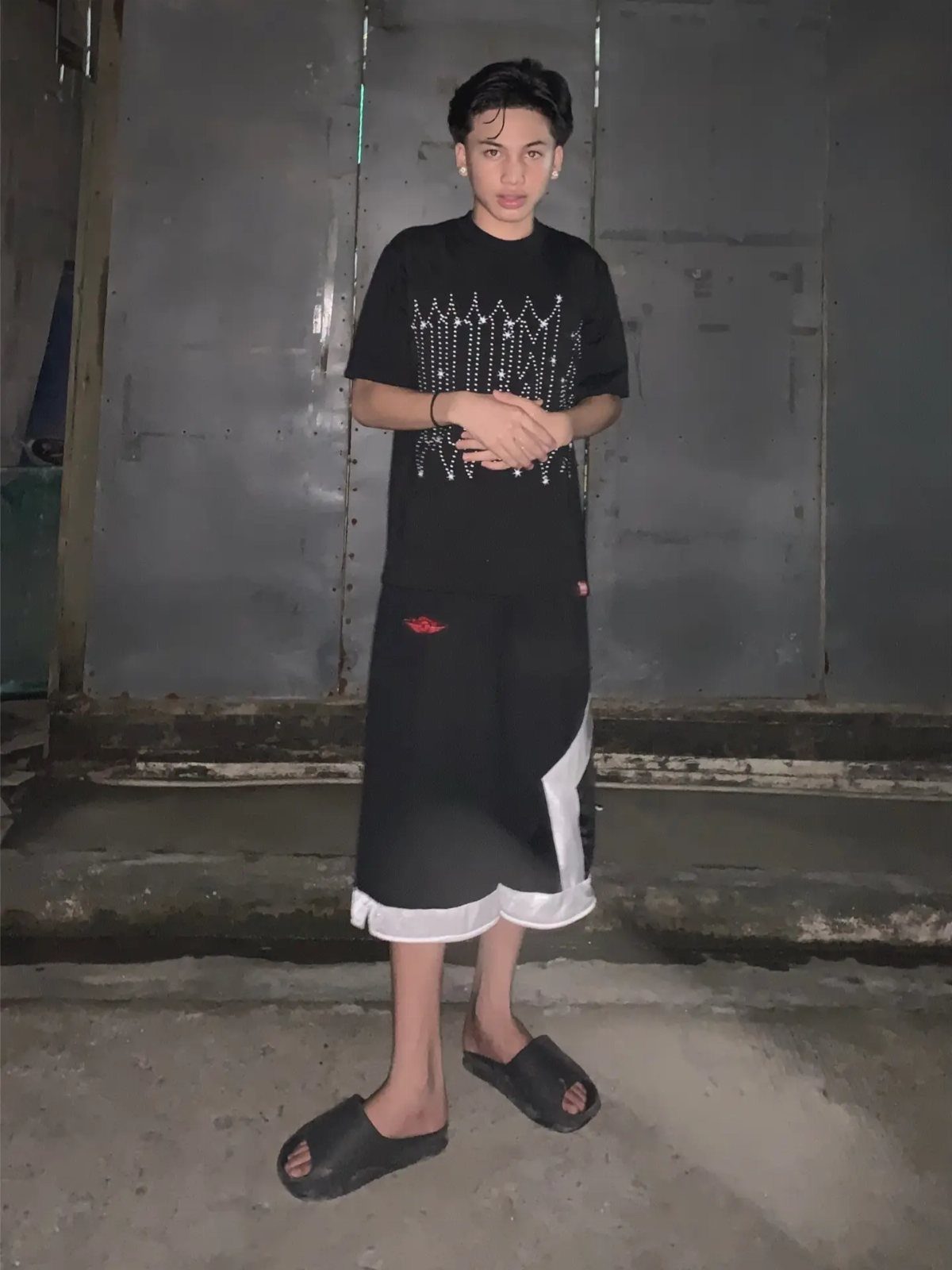 A young man posing in black graphic streetwear and shorts against a dark wall.