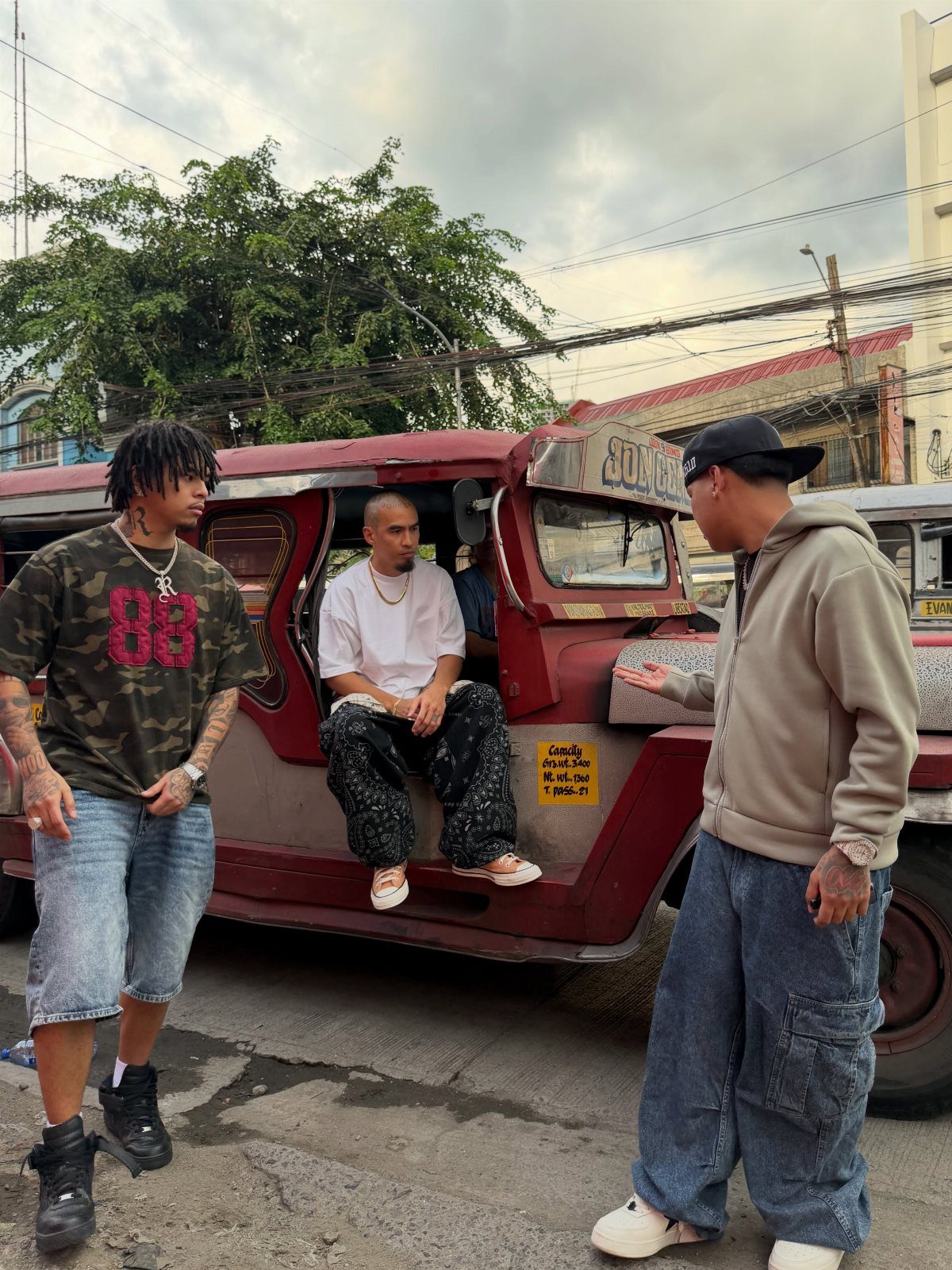 Three men wearing various streetwear styles posing next to a red jeepney on a street.