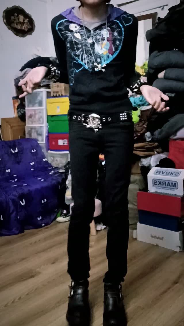 A person posing in a black graphic hoodie, studded belt, and black skinny jeans.