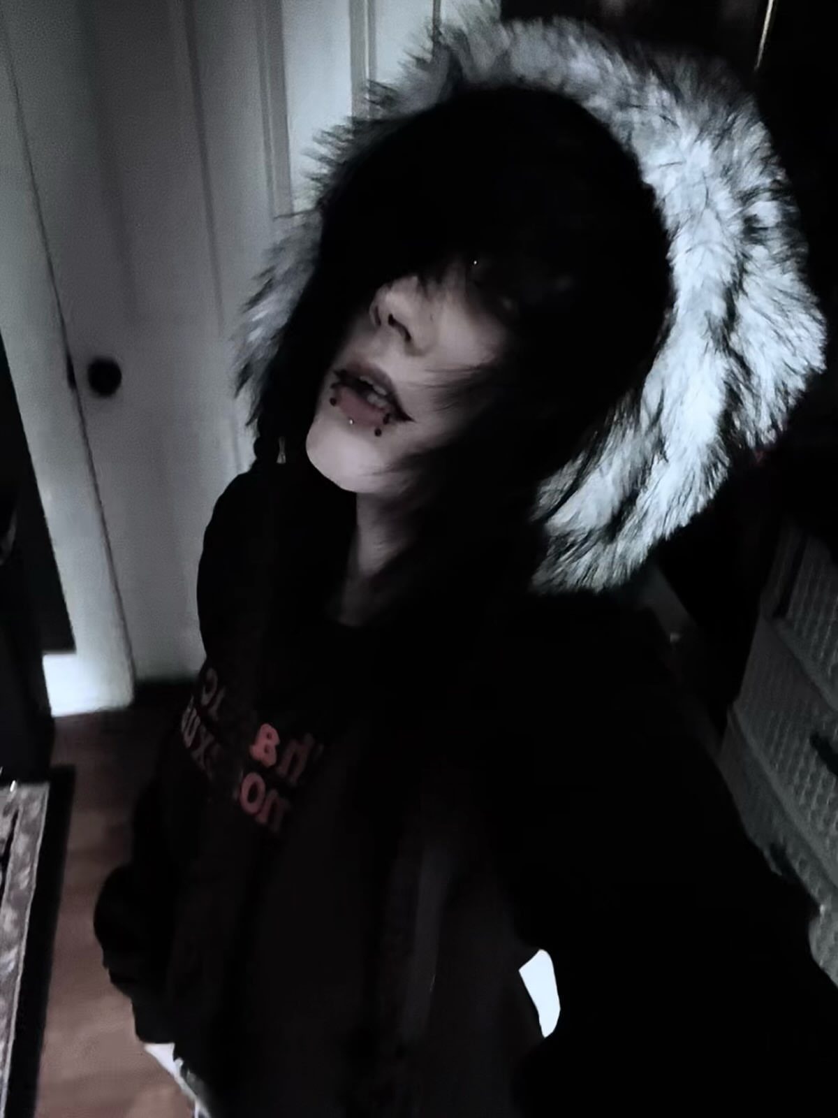 Person with dark hair and makeup wearing a black hoodie with grey faux-fur trim.