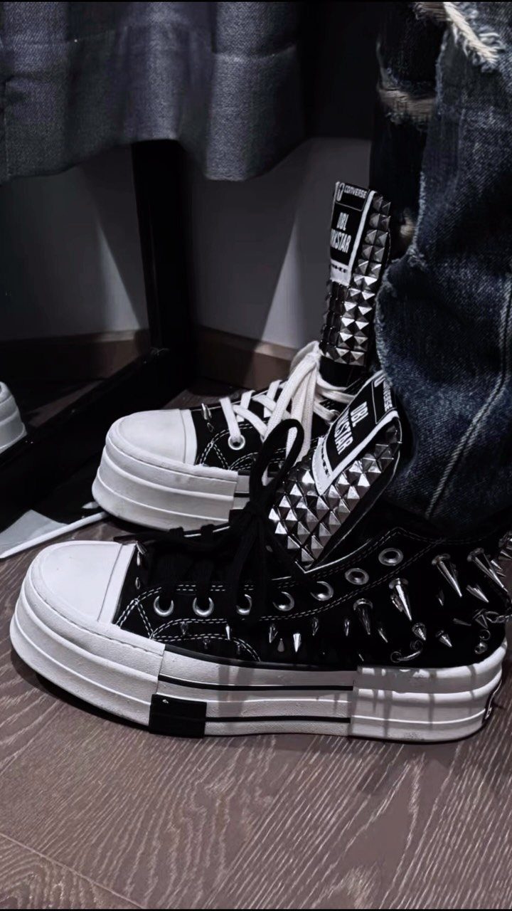 Black studded high-top platform sneakers worn with ripped denim jeans on a wooden floor.