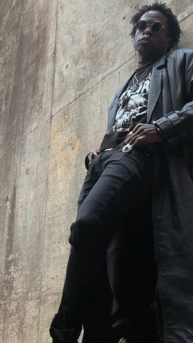 Man in a long black trench coat and sunglasses posing against a concrete wall.