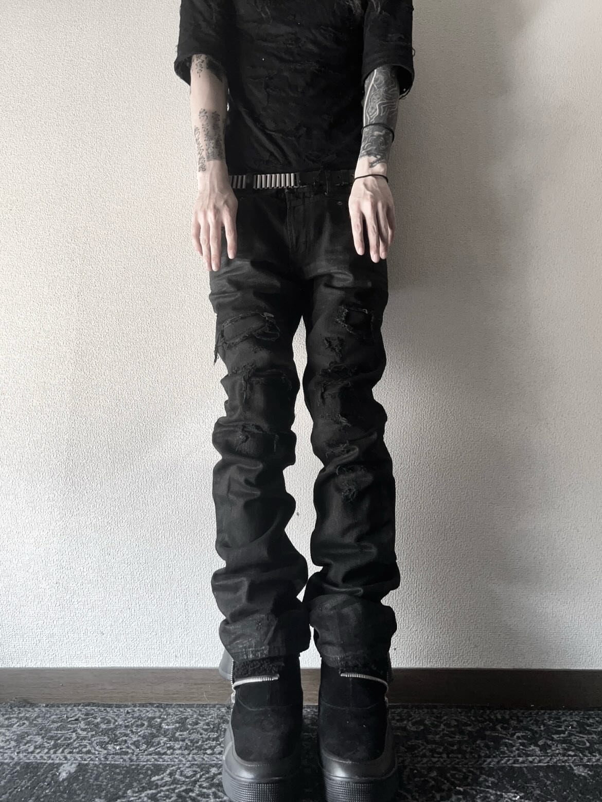 A person wearing black t-shirt, ripped black jeans, and platform boots standing against a wall.