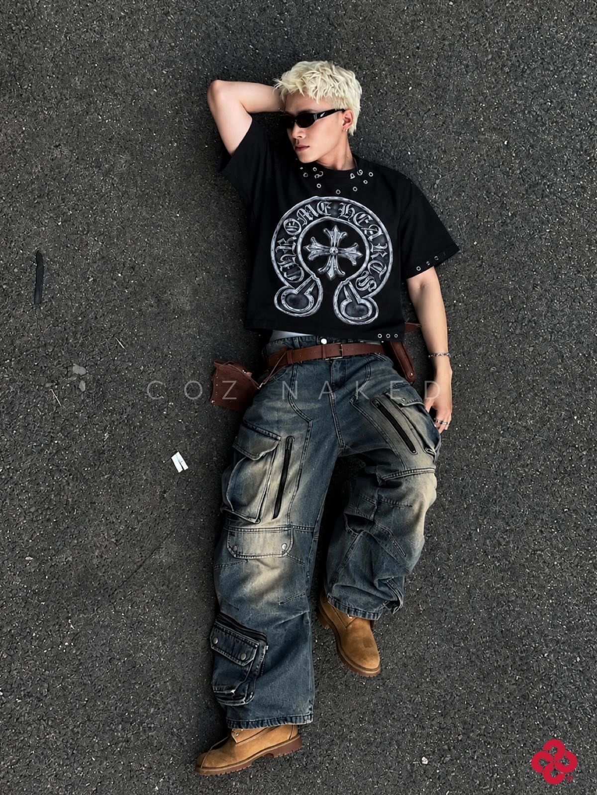A blonde person in grunge fashion lying on dark asphalt wearing baggy cargo pants and a graphic t-shirt.