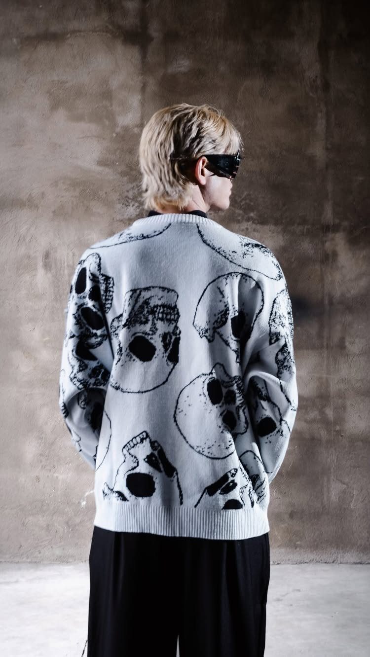 Person wearing an oversized light grey sweater with large black skull prints against a concrete wall.