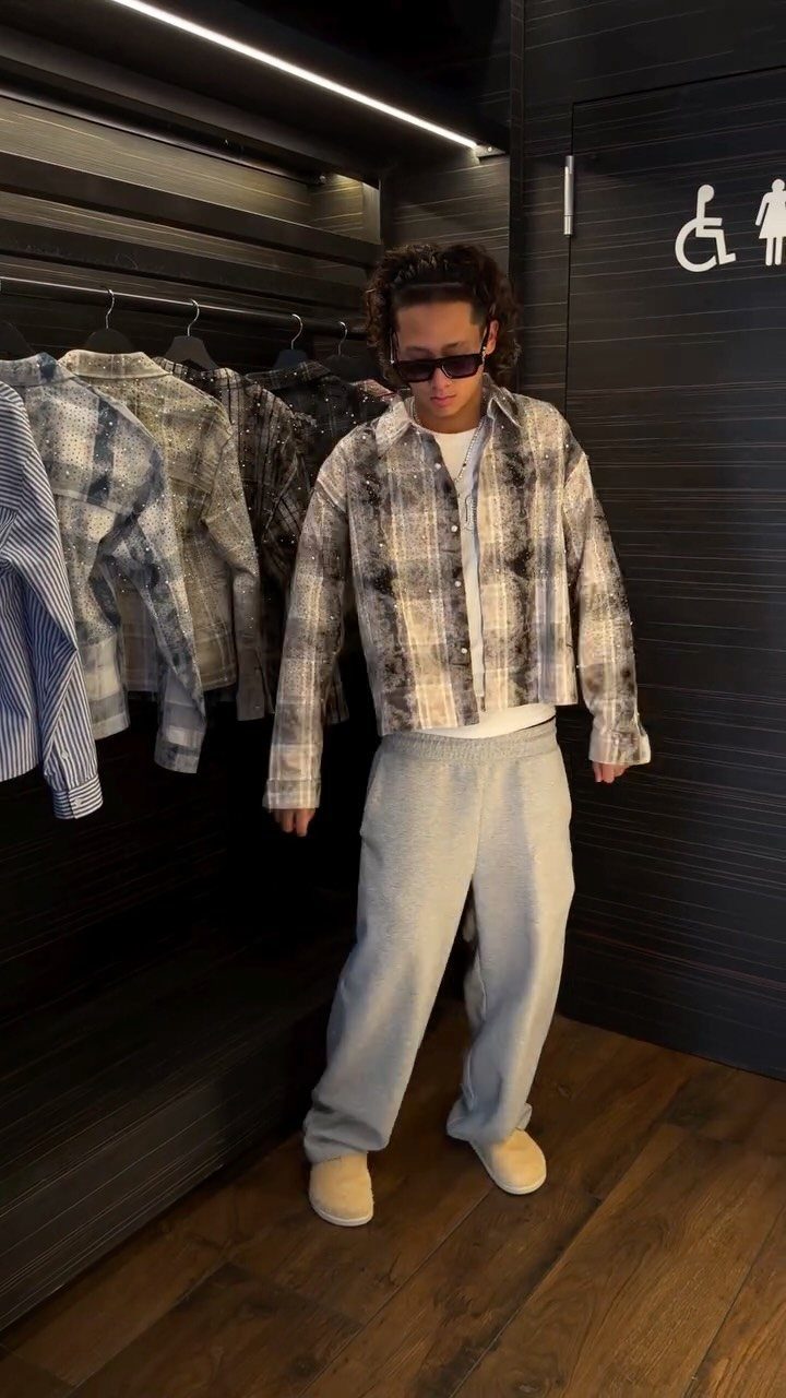 A person posing in a cropped plaid shirt, baggy sweatpants, and sunglasses in a dark room.