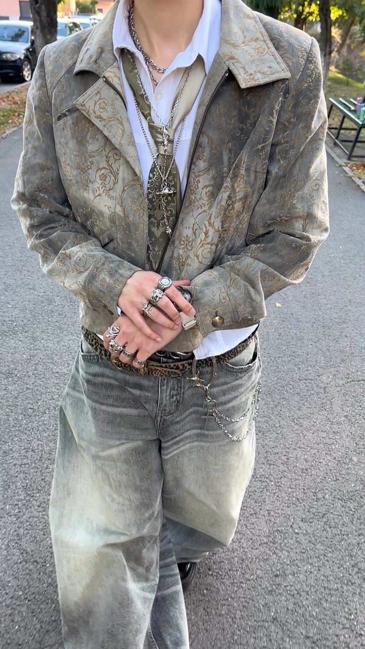 Person wearing a patterned grey jacket, silver jewelry, and baggy grey jeans standing on a street.