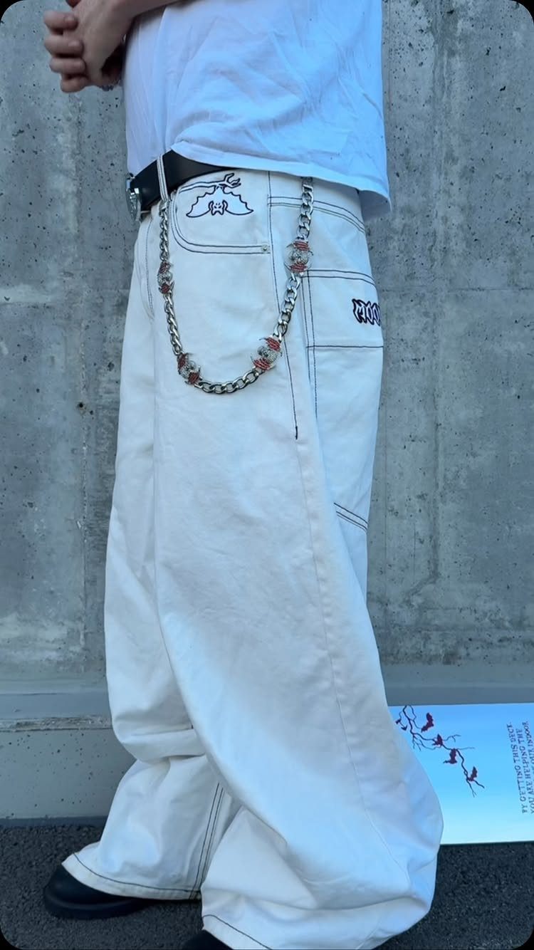 Person wearing baggy white denim pants with a silver chain and black belt against a concrete wall.