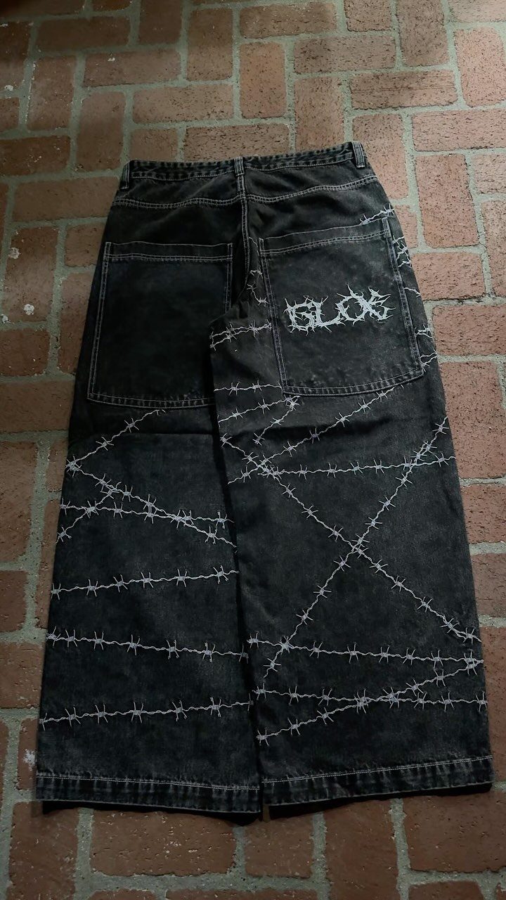 Black baggy denim pants featuring white barbed wire graphics lying on a brick floor.