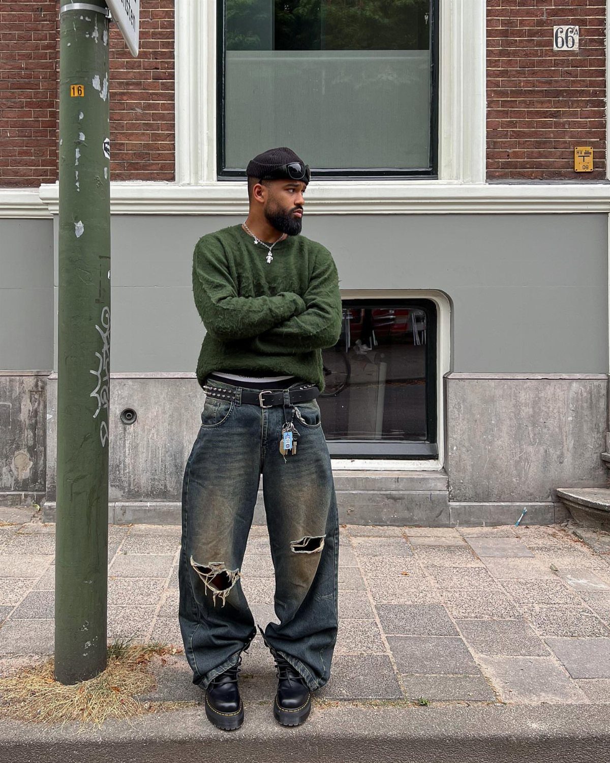 A man wearing a green sweater, ripped baggy jeans, and a black beanie standing on a sidewalk.