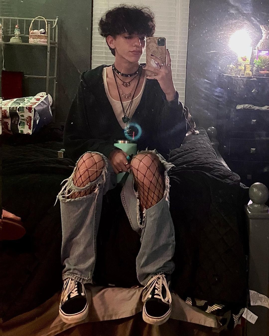 A person wearing ripped denim jeans with fishnet tights and layered necklaces sitting on a bed.