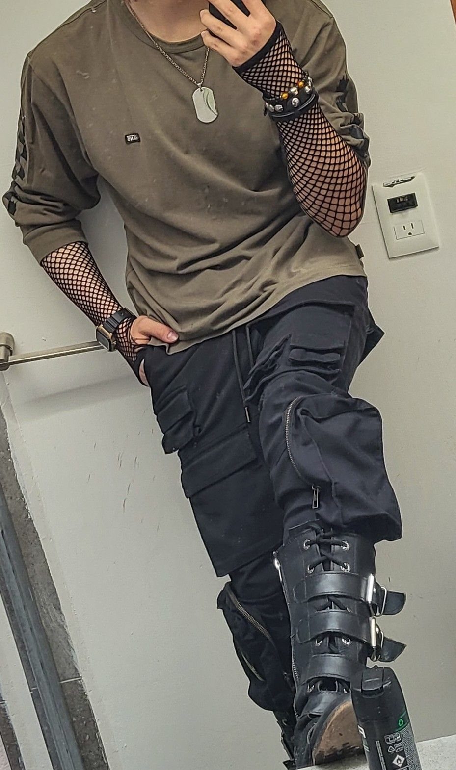 Person wearing olive green shirt, black cargo pants, buckled boots, and black fishnet sleeves.