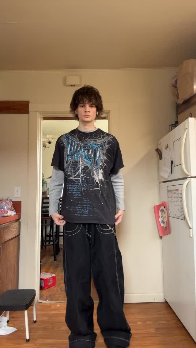 A person wearing a black graphic t-shirt, grey long-sleeve shirt, and baggy black trousers.