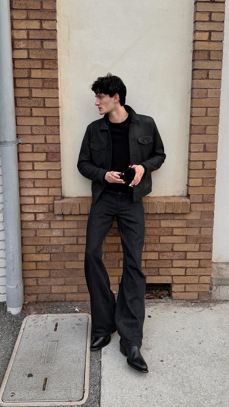 A person in an all-black outfit standing against a brick wall wearing black leather boots.