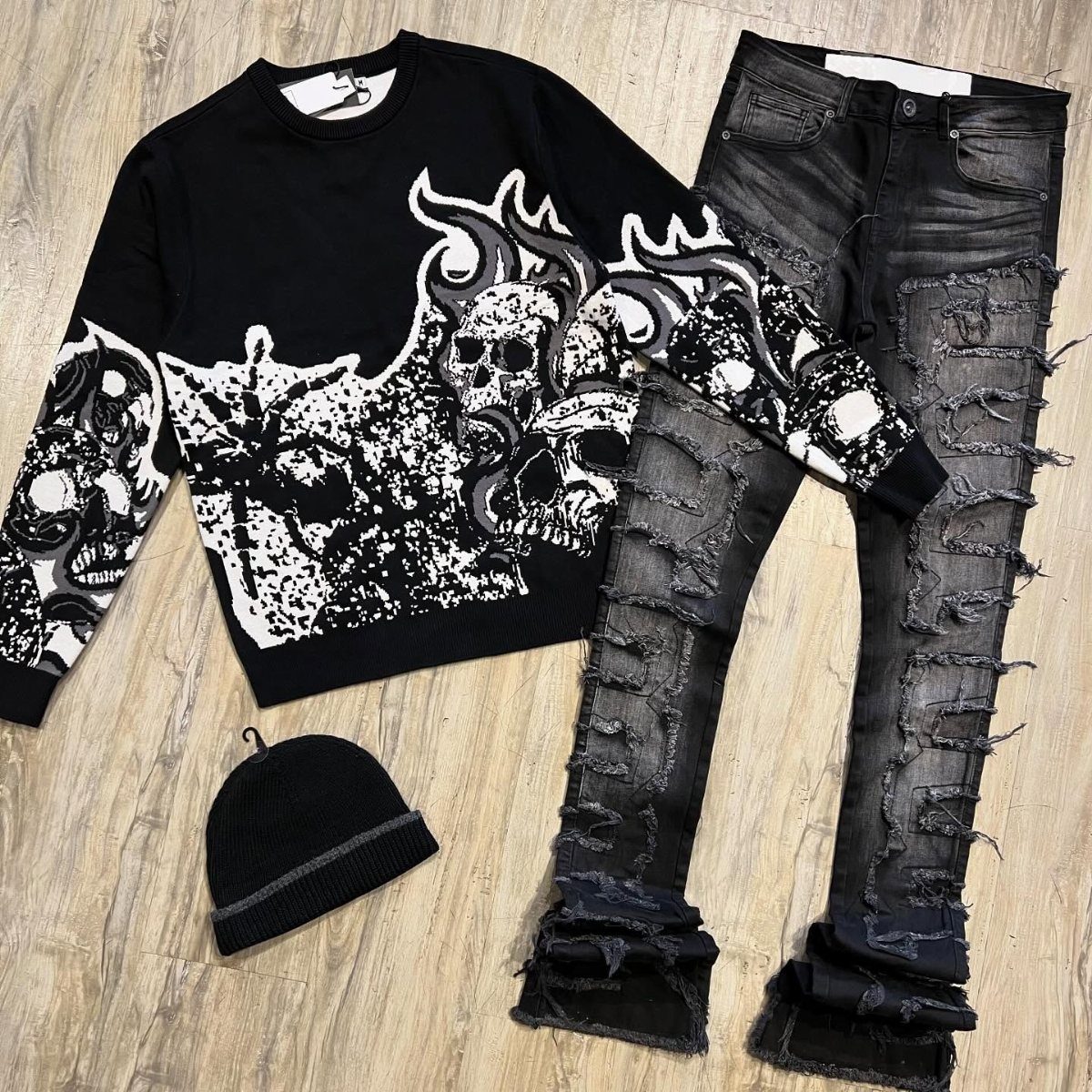 Ripped black denim jeans, a skull graphic sweatshirt, and a black beanie on a wooden floor.