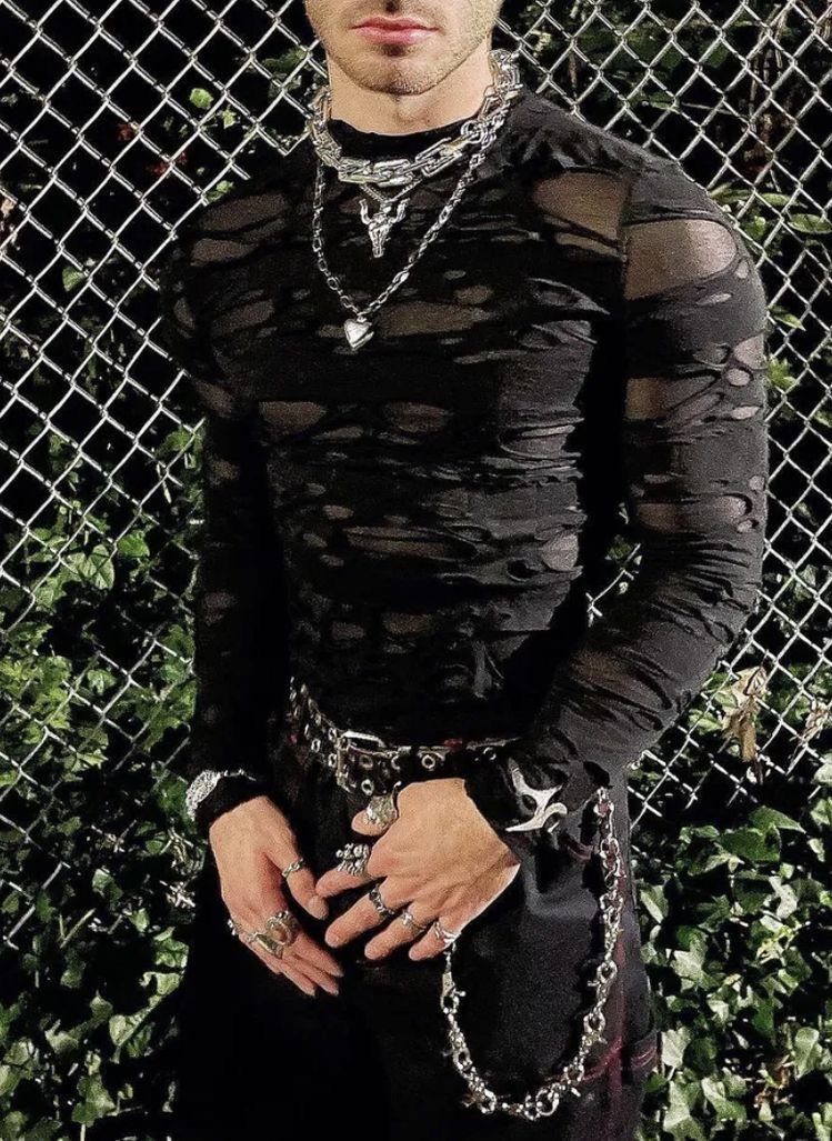 Person wearing distressed black clothing and layered silver chains standing before a chain-link fence.