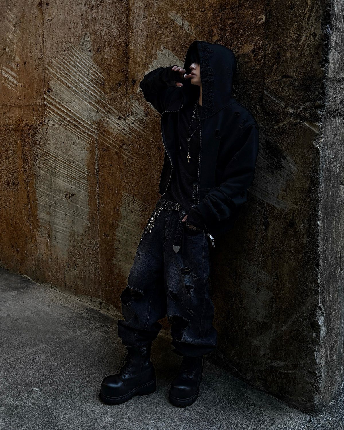 A person in a black hoodie and distressed black jeans leaning against a concrete wall.