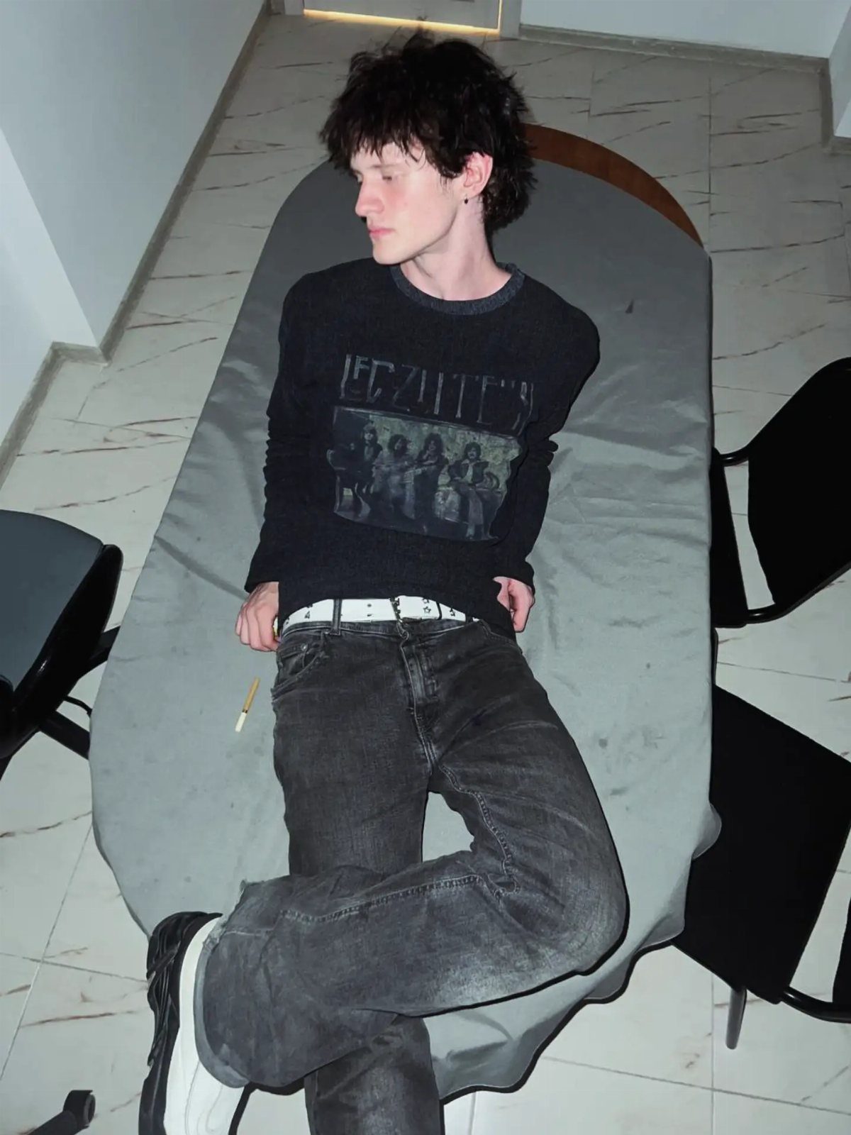 Young man with messy dark hair wearing a black graphic t-shirt and dark jeans lying on a table.