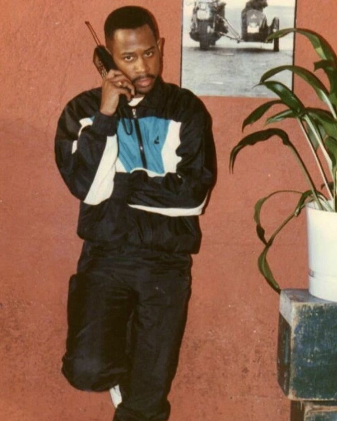 Man in a color-blocked tracksuit talking on a vintage mobile phone against a red wall.