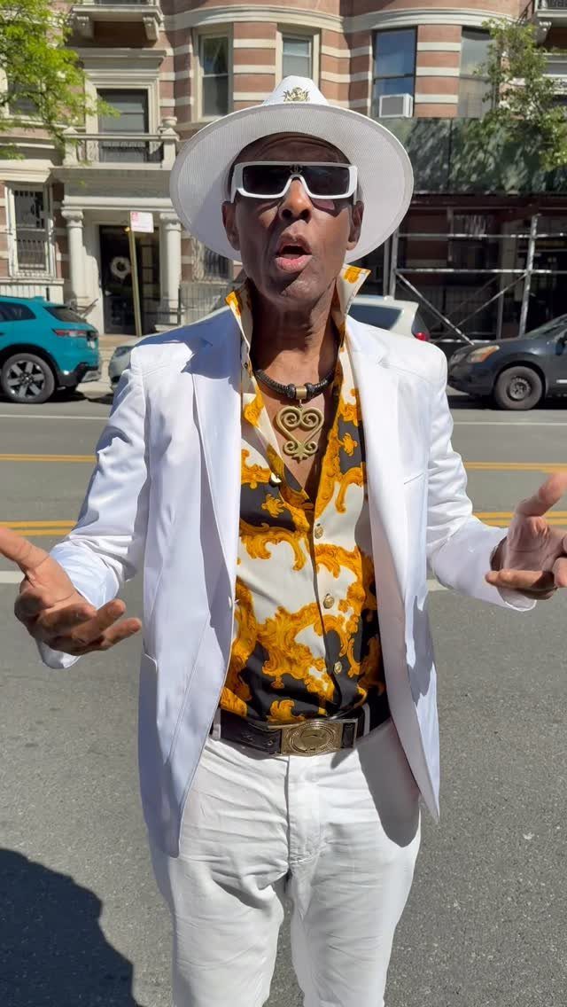 Man in white suit, patterned shirt, and large gold jewelry posing on a city street.